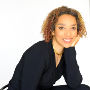 a multiracial Black woman with coffee-colored skin and short curly brown and golden hair, leans forward resting her cheek on her hand. She wears a black shirt and a gold necklace and smiles at the viewer. Photo by Beverlie Lord.