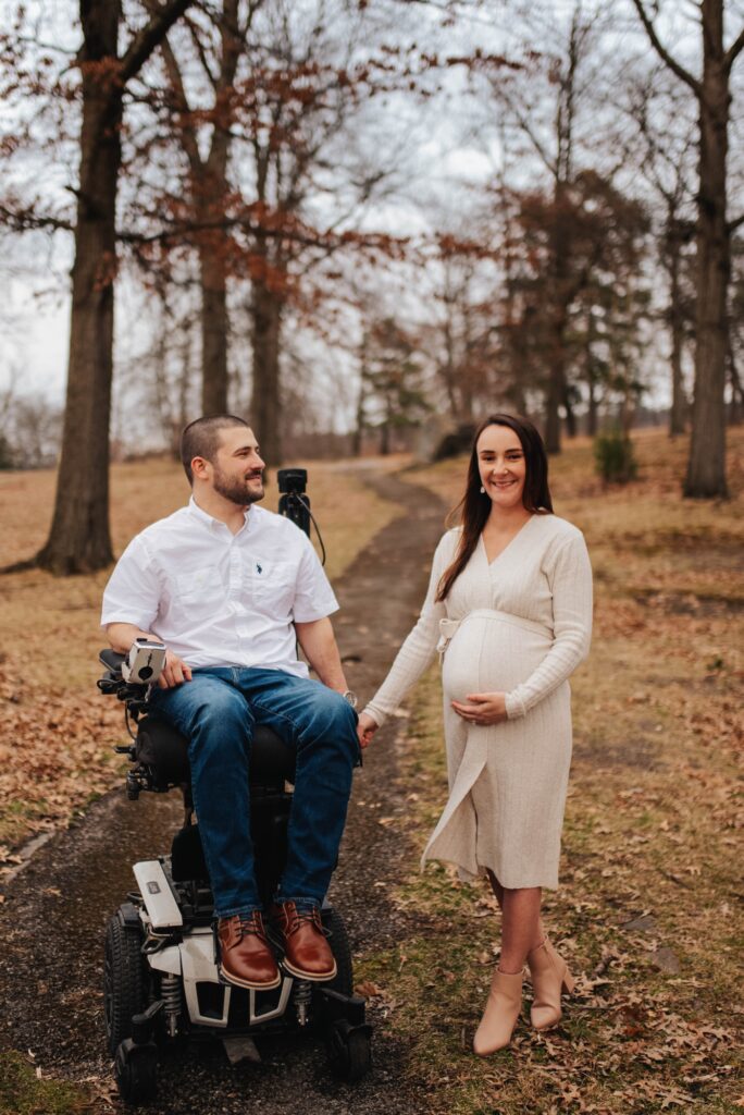 A man in a wheelchair looks adoringly at his pregnant wife. The couple is pictured in a Fall woodland setting