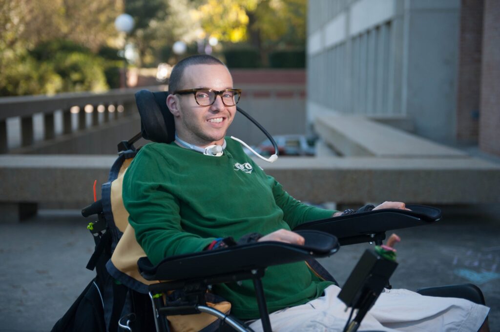 Smiling young man in a power chair on a college campus. He is wearing glasses and a green sweater. He appears to be happy and at peace.