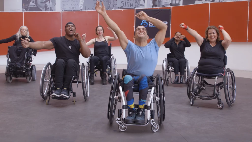 A group of wheelchair users wave their hands in the air during a high-energy fitness class