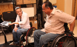 A smiling clinician in a wheelchair records information on a laptop as he chats with another man in a wheelchair.