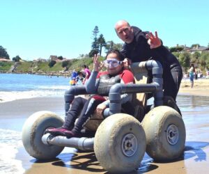 A smiling woman in an adaptive beach wheelchair, wearing a SCUBA outfit and goggles, gives the all is well sign with her right hand as a volunteer pushes her towards the ocean in Santa Cruz, California.