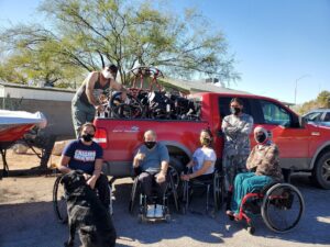 A group of facemask-wearing people in wheelchairs sit in front of a red pick-up truck. loaded with wheelchairs for those in need