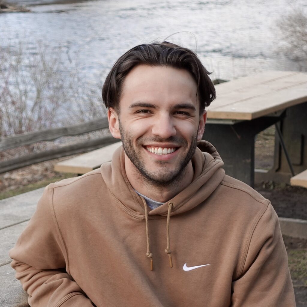 A man with short brown hair and a neatly trimmed beard, smiling warmly while wearing a tan hoodie. He is sitting outdoors near a picnic table, with a calm river and bare trees in the background.