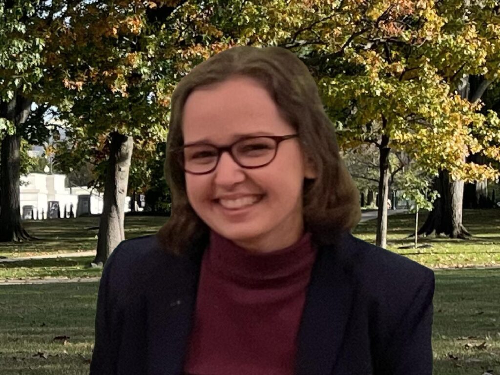 A smiling woman with short brown hair and glasses, sitting outdoors in a park with trees and autumn leaves in the background. She is wearing a dark blazer and a burgundy turtleneck.