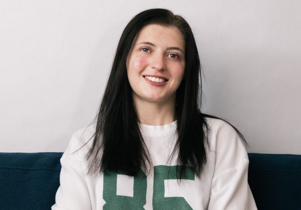 A woman with straight dark hair, smiling warmly while seated on a blue couch. She is wearing a white sweater with the number '85' in green on the front. The background is plain and light-colored.