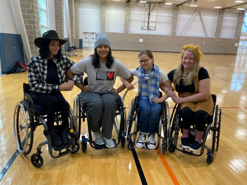 Four students in wheelchairs, all dressed casually for a Fall event, link arms on a basketball court