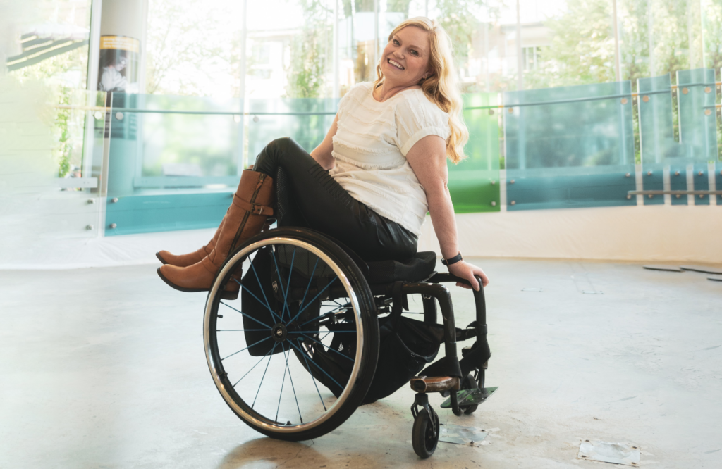 A smiling blonde woman sits sideways on a wheelchair as she shows off new accessible fashion items, including knee-length brown boots