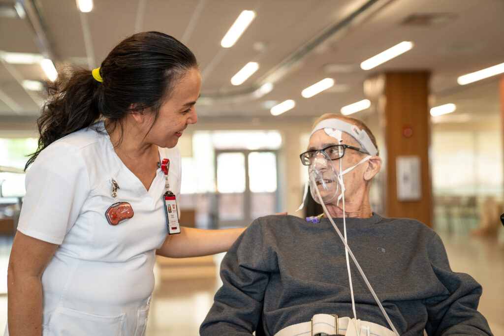 A nurse attends to a patient at the Craig H. Neilsen Rehabilitation Hospital in Utah. The female nurse smiles as she places a comforting hand on the male patient's shoulder. He is wearing a headband and a clear mask to aid his breathing.