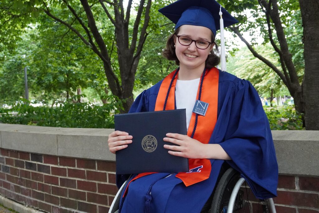 Smiling University of Illinois, Urbana-Champaign student on graduation day. Sitting in a wheelchair and posing with her diploma, the student wears spectacles, a cap, a navy blue gown over a white T-shirt, and an orange sash