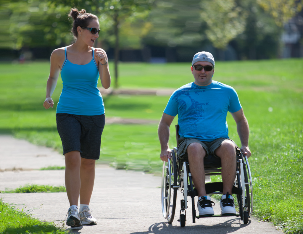 individual walking with individual using a wheelchair through a park setting