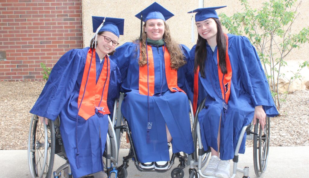 Three women in wheelchairs pose together after graduation, wearing blue graduation robes.