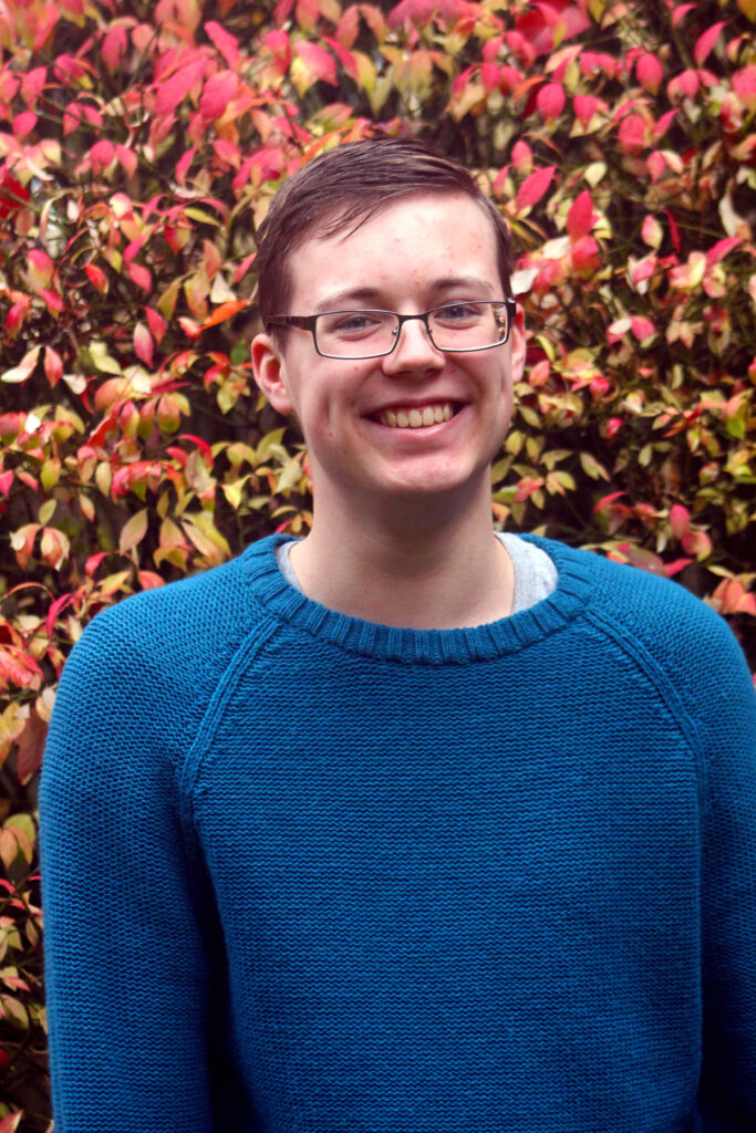 A young man with short brown hair and glasses, smiling warmly while wearing a blue sweater. He is outdoors in front of vibrant bushes with red and green leaves.