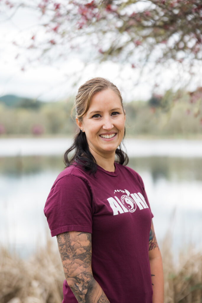 A smiling woman with light brown hair pulled back, wearing a maroon t-shirt with the word 'Aloha' on the front. She is standing outdoors by a lake, with trees and branches in bloom in the background.