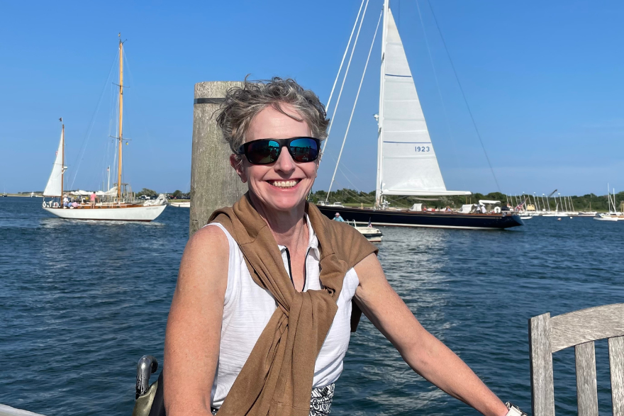 Woman in a wheelchair wearing sunglasses and a brown sweater over her shoulders smiles in front of a harbor with sailboats under a clear blue sky.