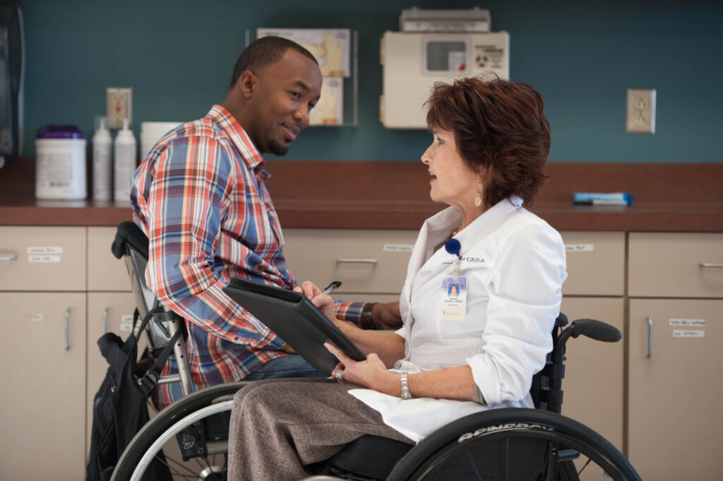 A man in a wheelchair speaking with a healthcare professional, also in a wheelchair, in a medical office.