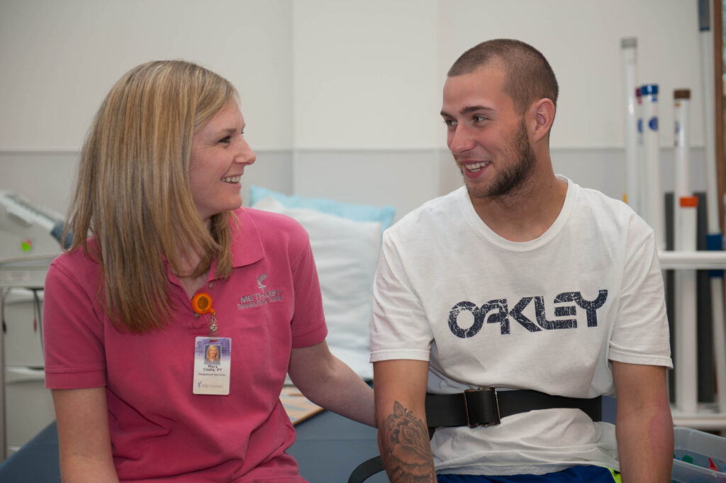 A therapist in a pink shirt smiles at a young man in a white t-shirt, seated in a rehab setting with equipment in the background.