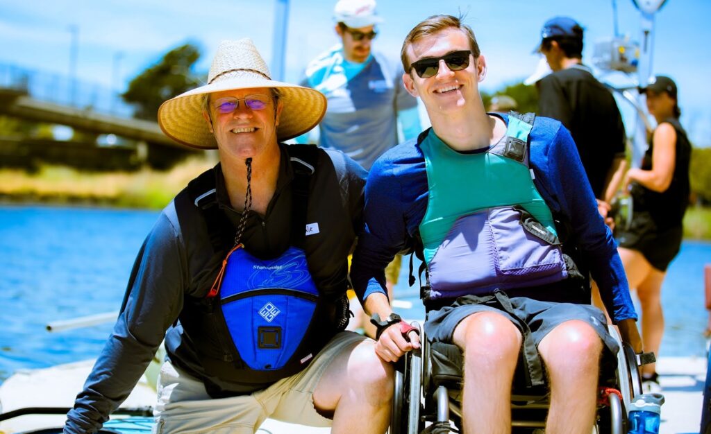 Two individuals, one wearing a straw hat and the other in a wheelchair with a life vest, smiling together near a waterfront. A group of people and water activity equipment are visible in the background under a bright blue sky.