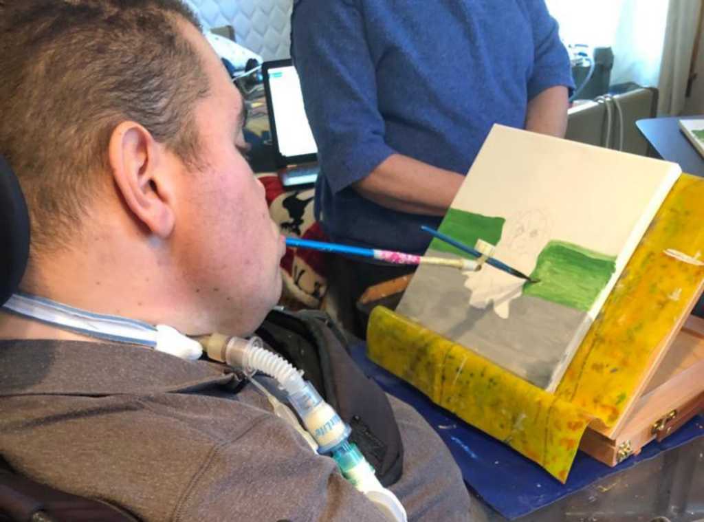 A man using a ventilator paints with a brush held in his mouth. He is working on a canvas positioned on a tabletop easel, with green and gray colors partially filled in. Another person stands nearby, observing.