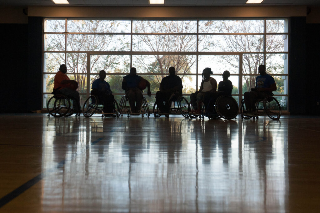 A group of people in wheelchairs sits in a gym facing large windows, silhouetted against the natural light.
