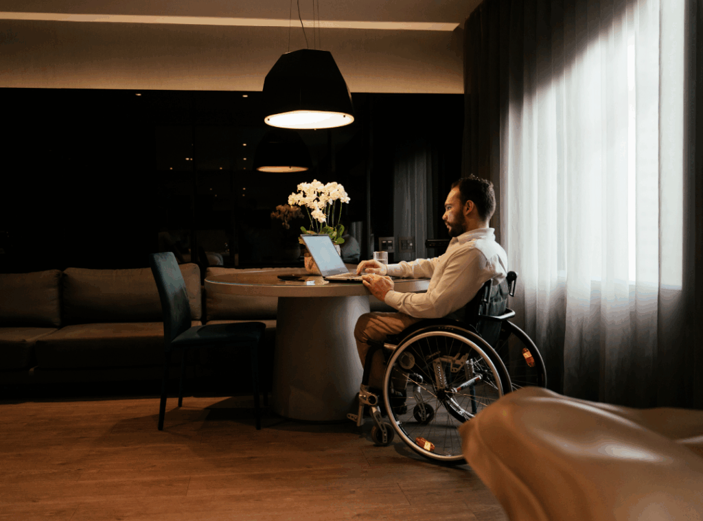 A man in a wheelchair with dark hair, a light shirt, and dark pants sits in the lobby of a hotel, using a laptop.