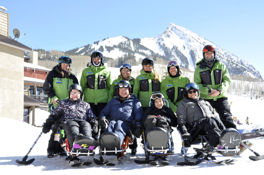 Adaptive skiers and instructors pose together on a snowy slope at the Adaptive Sports Center of Crested Butte, with three mono-skiers in front and instructors standing behind them against a mountain backdrop.