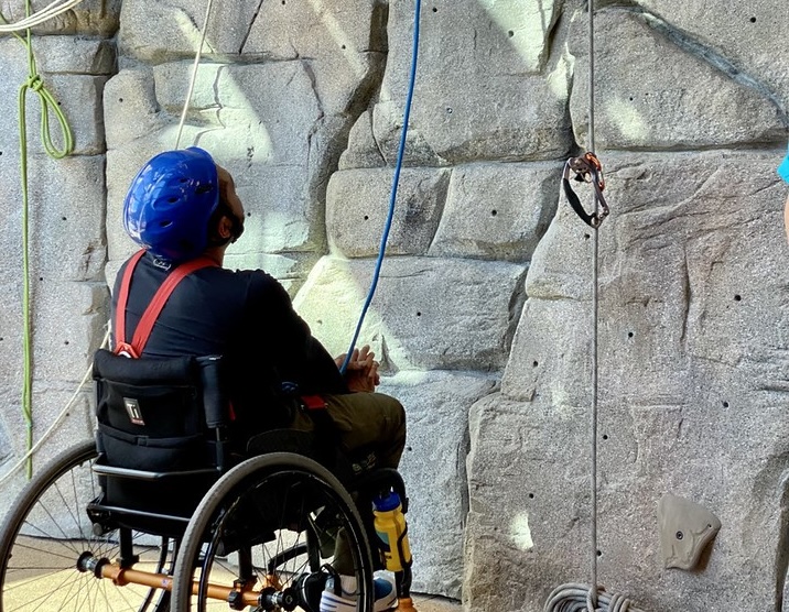 A person using a wheelchair, wearing a helmet and harness, faces an indoor rock climbing wall with ropes and climbing holds, preparing for an adaptive climbing activity.