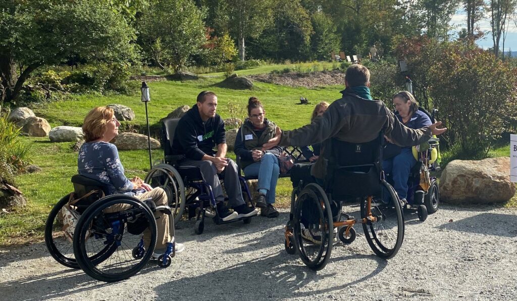Group of people, some in wheelchairs, some sitting on a bench, talking with each other in a green park area.