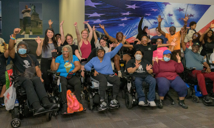 A large, diverse group of people, many using wheelchairs, sit and stand together indoors, smiling and raising their hands in celebration in front of a large American flag backdrop.