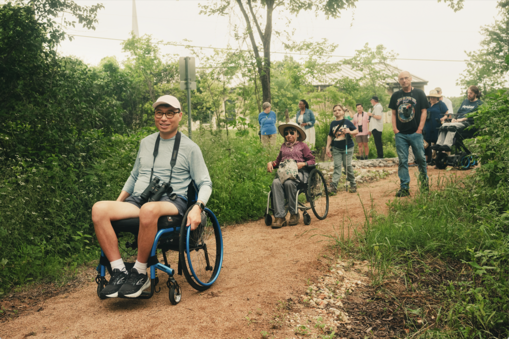 A group of people of varying ages, including several wheelchair users, travel together along a tree-lined gravel path in a park; a smiling person in a wheelchair with binoculars leads the group.