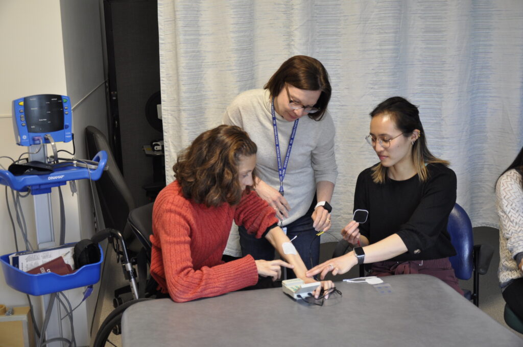 Three people sit around a table in a clinical lab setting, learning to use rehabilitation equipment with electrodes attached to one person’s arm.