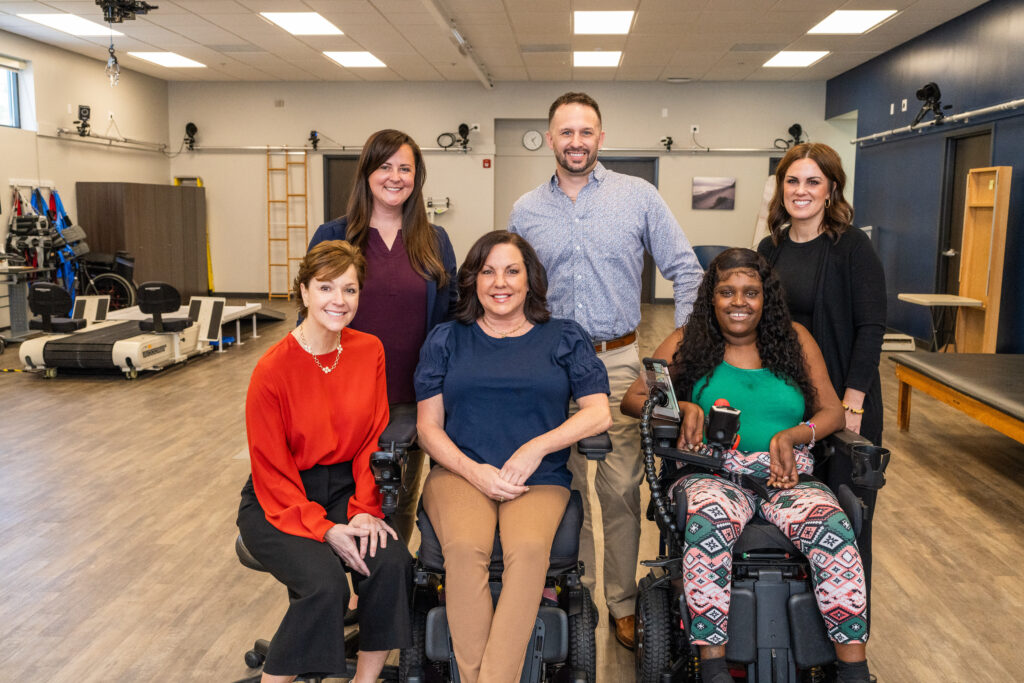 Six people pose in a rehabilitation therapy room, including two women seated in power wheelchairs and three people standing behind them.
