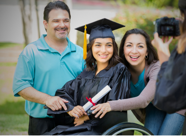 Young woman in a wheelchair wearing a graduation cap and gown poses with her parents while holding her diploma.