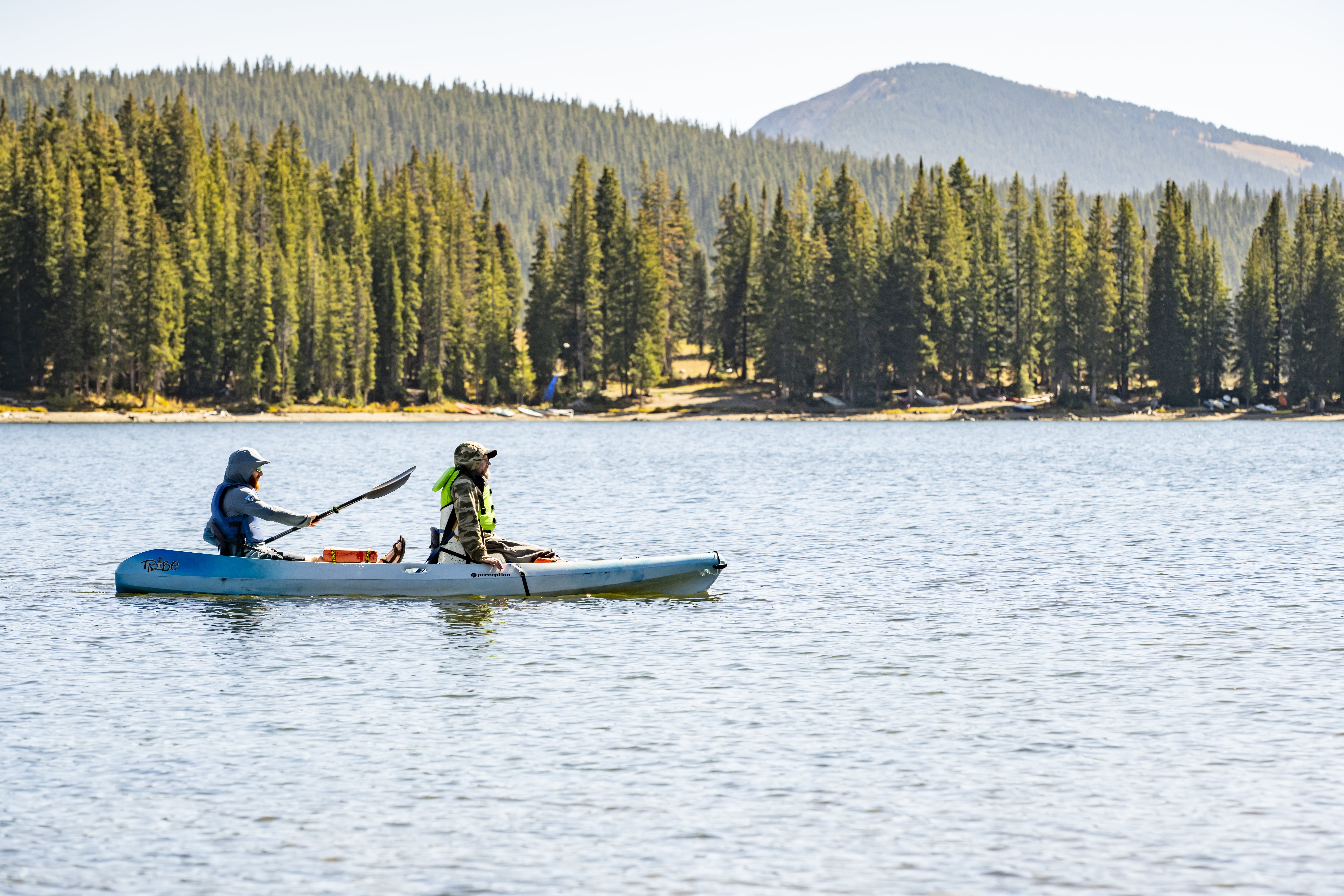 Two people paddle a blue tandem kayak across a calm mountain lake surrounded by evergreen forests and mountains.